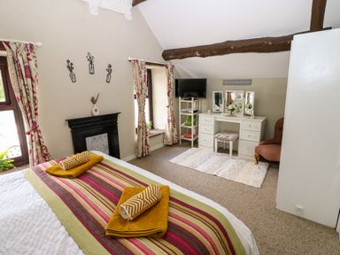 A bedroom with a bed featuring striped bedding and folded towels a white vanity table with mirrors a chair a TV on a stand and a fireplace at River Cottage in Eamont Bridge