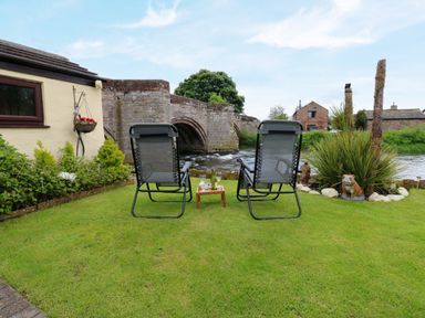 A garden with two chairs and a small table by a river and stone bridge at River Cottage in Eamont Bridge