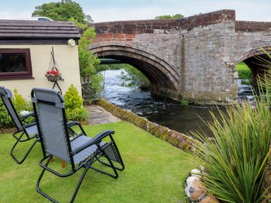 A garden with green lawn and two chairs next to a stone bridge over a river at River Cottage in Eamont Bridge