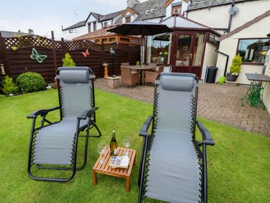 A garden with two reclining chairs a small wooden table with a bottle and two glasses and a patio with table and chairs at River Cottage in Eamont Bridge