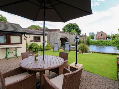 A patio with a round table and chairs under an umbrella next to a garden with chairs and a river by a stone bridge at River Cottage in Eamont Bridge
