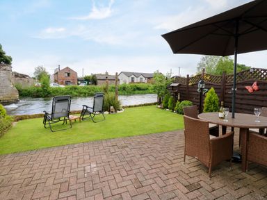 A garden with two reclining chairs and table near a river and a patio with a table umbrella and chairs at River Cottage in Eamont Bridge