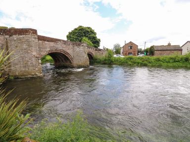 A stone bridge over a river with houses and greenery in the background at River Cottage in Eamont Bridge