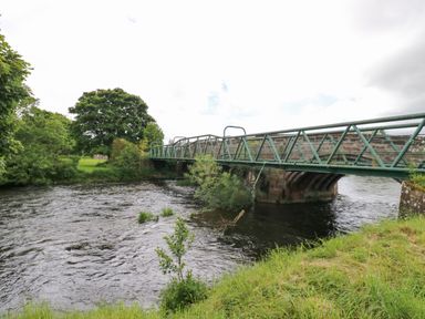 A metal pedestrian bridge over a river with trees and grass on the riverbanks at River Cottage in Eamont Bridge