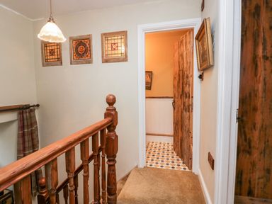 A hallway with wooden banister stairs a light fixture three framed stained glass windows and an open door showing a tiled floor at Eden Vale in Broughton-In-Furness