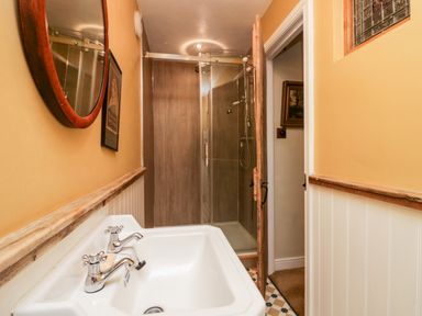 A bathroom with a white sink and taps a round mirror a framed picture a shower with glass doors and a wooden door at Eden Vale in Broughton-In-Furness