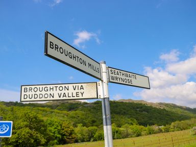 A road sign showing directions to Broughton Mills Seathwaite Wrynose and Broughton via Duddon Valley with hills and trees in the background at Eden Vale in Broughton-In-Furness