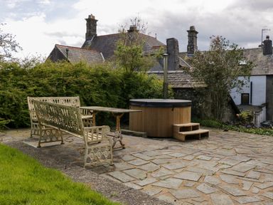 An outdoor patio with metal benches a table and a wooden hot tub surrounded by bushes and buildings at Eden Vale in Broughton-In-Furness