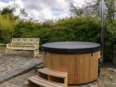 An outdoor area with a wooden hot tub with a cover and a vintage metal bench on a stone patio surrounded by greenery at Eden Vale in Broughton-In-Furness