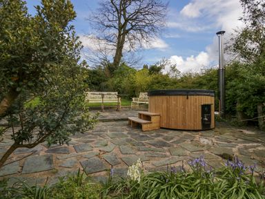 A garden patio with a wooden hot tub wooden steps and white metal benches surrounded by greenery at Eden Vale in Broughton-In-Furness