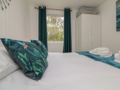 A bedroom with a bed with floral pillows folded towels on the bed a window with dark curtains and a white wardrobe at Cassia Kilkhampton
