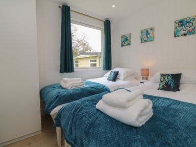 A bedroom with two single beds with blue throws and folded white towels by a window with blue curtains at Cassia Kilkhampton