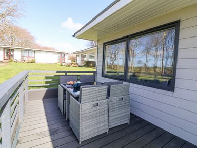 An outdoor deck with a glass table and six wicker chairs next to a house with large windows at Cassia in Kilkhampton
