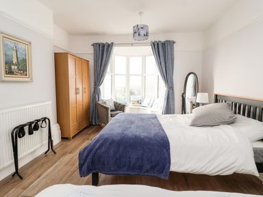 A bedroom with a bed covered with white and blue bedding a wooden wardrobe gray curtains and a window seat at 46 The Promenade in Withernsea