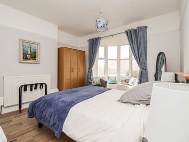 A bedroom with a bed, blue blanket, wooden wardrobe, bay window with curtains, cushions and an armchair at 46 The Promenade in Withernsea