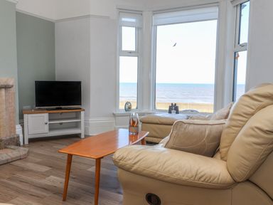A living room with a TV on a stand a wooden coffee table and beige leather sofas next to large windows overlooking the sea at 46 The Promenade in Withernsea