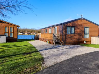 A gravel driveway leading to a brown wooden lodge near a lake with other lodges visible in the background at Gressingham 7 in Warton near Carnforth