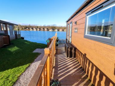 A wooden deck with railing next to a building by a lake with houses across the water at Gressingham 7 Warton near Carnforth