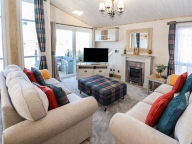A living room with two beige sofas a plaid ottoman a fireplace a TV and large windows with curtains at Gressingham 7 in Warton near Carnforth