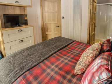 A bedroom with a bed covered in red and gray plaid bedding two wooden doors a chest of drawers with a television and an open door leading to a shower at Gressingham 7 in Warton near Carnforth