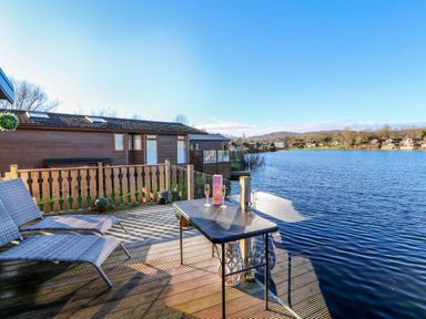 A lakeside wooden deck with two lounge chairs and a table with glasses and a box at Gressingham 7 in Warton near Carnforth