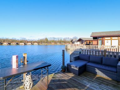 A deck with wicker seating and a table overlooking a lake with wooden cabins in the background at Gressingham 7 in Warton near Carnforth