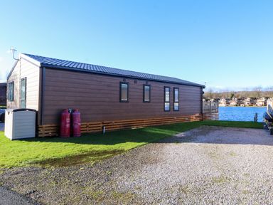 A brown mobile home with windows and gas cylinders next to a gravel driveway by a lake at Gressingham 7 in Warton near Carnforth