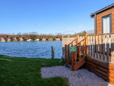 Lake with wooden deck and stairs in the foreground and row of cabins across the water at Gressingham 7 in Warton near Carnforth