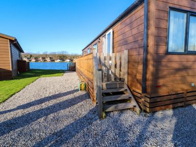 A wooden cabin with steps and railing beside a gravel pathway next to a lake at Gressingham 7 in Warton near Carnforth