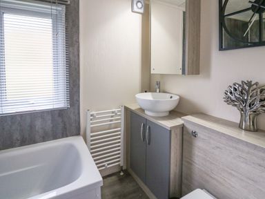 A bathroom with a bathtub, sink on a cabinet, a wall mirror, a towel radiator, and a decorative tree sculpture at 43 The Deri in Llanarth