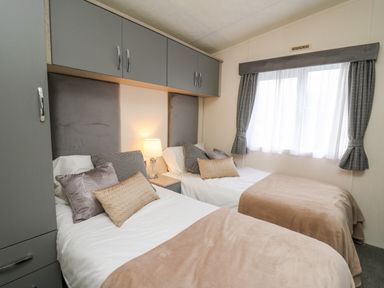 A bedroom with two single beds alongside a window with curtains and overhead storage cabinets at 43 The Deri in Llanarth
