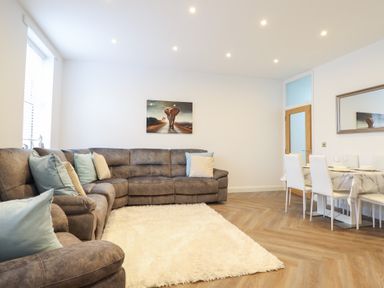 A living room with a grey sectional sofa, blue and beige pillows, a white fluffy rug, a dining table with white chairs, and an elephant painting on the wall at Gwydyr Bach 1 in Llanrwst