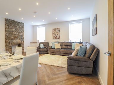 A living room with a stone fireplace a brown sectional sofa with pillows a white rug two windows and a dining table with white chairs at Gwydyr Bach 1 in Llanrwst