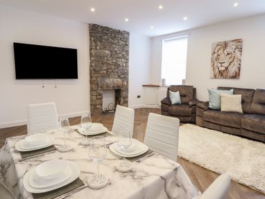 A dining area with a marble table and white chairs in an open living room with brown sofas and a stone fireplace at Gwydyr Bach 1 in Llanrwst