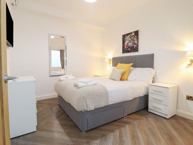 A bedroom with a grey upholstered bed two bedside tables a chest of drawers a wall mirror and a leopard painting at Gwydyr Bach 1 in Llanrwst