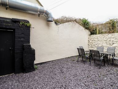 An outdoor patio area with a glass table and six chairs on a gravel ground with cream and stone walls at Gwydyr Bach 1 in Llanrwst