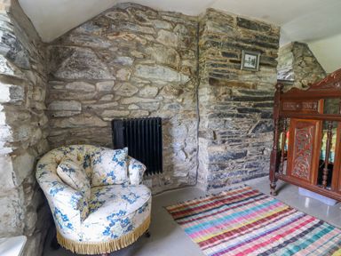 A corner with a floral armchair, black radiator, colorful striped rug, wooden carved furniture, and stone walls at Nyth Yr Eryr in Blaenau Ffestiniog