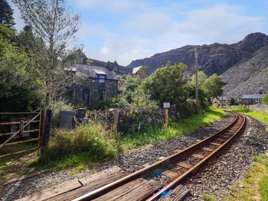 A curved railway track passing beside stone houses and trees in a rural area with mountains in the background at Nyth Yr Eryr in Blaenau Ffestiniog