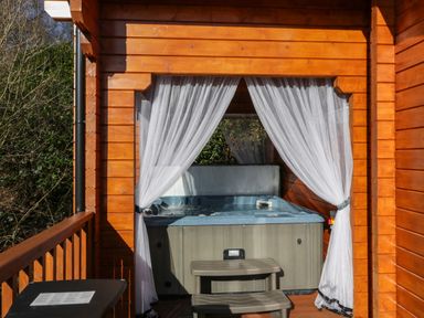 An outdoor hot tub on a wooden porch with sheer white curtains tied back at Little Lodge Balminnoch near Glenluce
