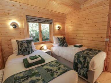 A bedroom with two single beds green patterned pillows and blankets a nightstand with a lamp and wooden walls at Little Lodge in Balminnoch near Glenluce