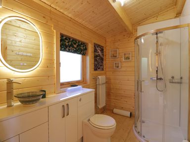 A bathroom with wooden walls a round illuminated mirror a vessel sink a toilet and a curved glass shower at Little Lodge in Balminnoch near Glenluce