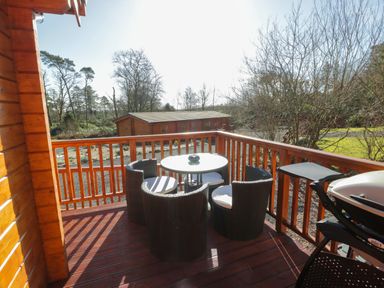 A wooden deck with a round table and four chairs and a barbecue grill with a wooden cabin and trees in the background at Little Lodge in Balminnoch near Glenluce