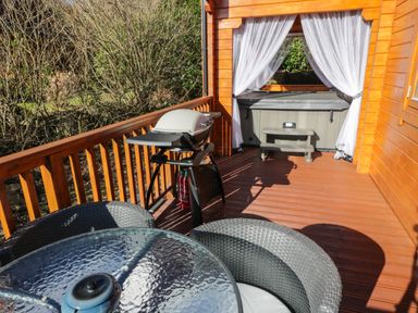 A wooden deck with a barbecue grill glass table with chairs and a hot tub with white curtains at Little Lodge in Balminnoch near Glenluce