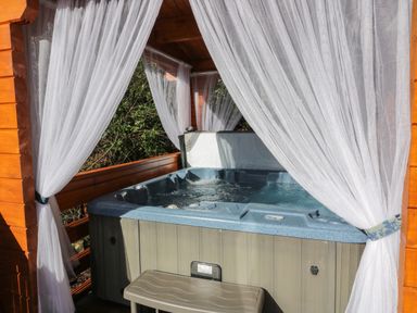 An outdoor hot tub with water jets under a wooden gazebo with white curtains tied back at Little Lodge Balminnoch near Glenluce