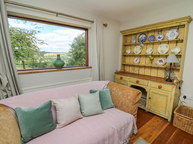 A living room with a sofa near a window and a yellow wooden cabinet with plates at Oak Apple Cottage in Presteigne