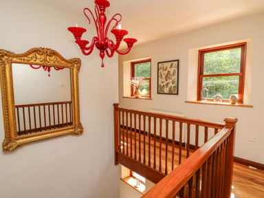 A wooden staircase landing with two windows decorated with glass items a large ornate gold mirror on one wall and a red chandelier hanging from the ceiling at Oak Apple Cottage in Presteigne