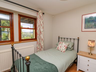 A bedroom with a single bed next to a white nightstand with a lamp and two windows with floral curtains at Oak Apple Cottage in Presteigne