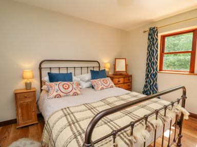 A bedroom with a metal bed frame bedside tables lamps a wooden dresser with a mirror and a window with patterned curtains at Oak Apple Cottage in Presteigne