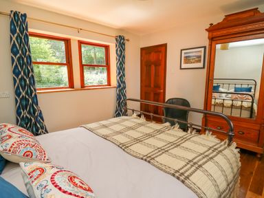 A bedroom with a metal bed frame a wooden wardrobe with mirror a chair and windows with blue curtains at Oak Apple Cottage in Presteigne