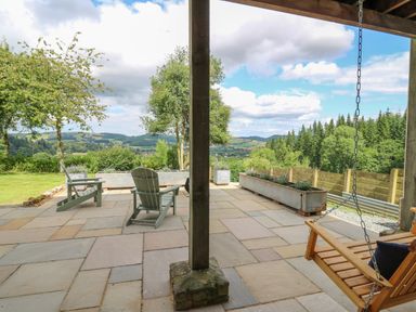 An outdoor patio with two green chairs a wooden swing and planters overlooking a landscape of trees and hills at Oak Apple Cottage in Presteigne